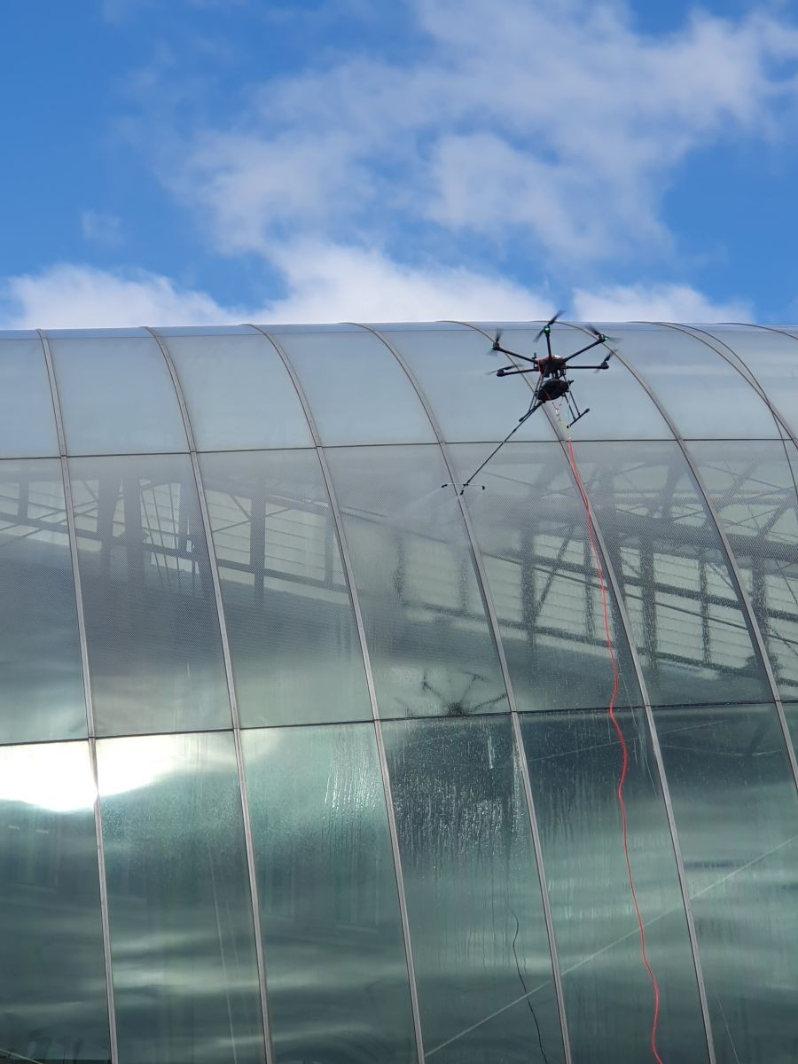 Tethered Drone Performing Glass Roof Inspection and Maintenance Over Modern Curved Building Under Blue Sky